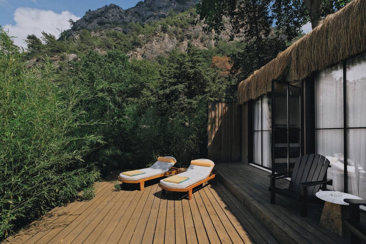 A wooden deck with two sun loungers and a table, a chair, and a thatched-roof building with large windows.