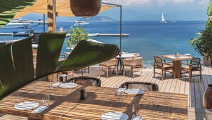 Une terrasse en bois à l’extérieur, avec des tables et des chaises, d’où l’on peut admirer une mer bleue et limpide parsemée de bateaux.