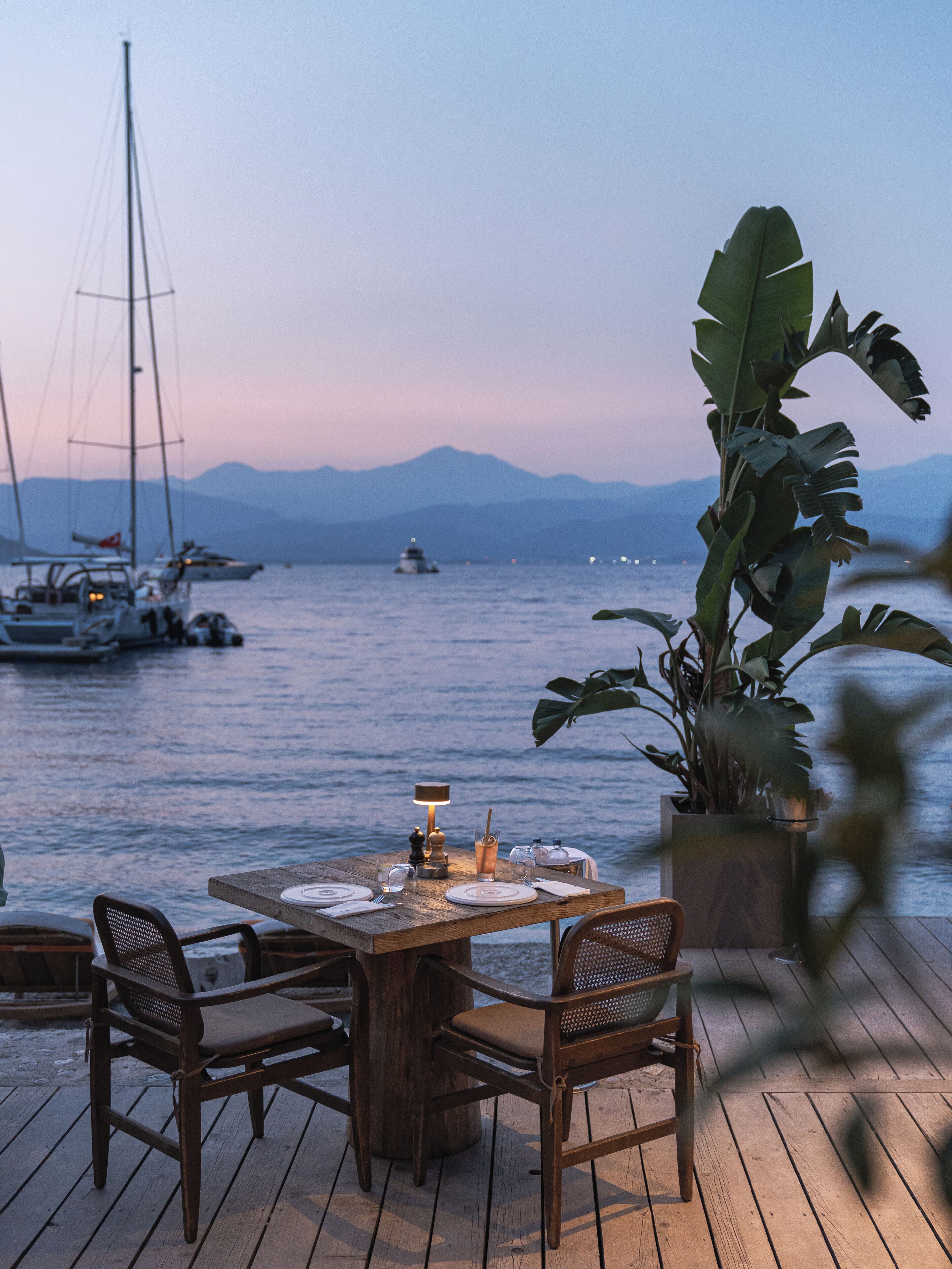 A wooden deck with a table set for dining, overlooking a calm body of water with boats and mountains in the background.