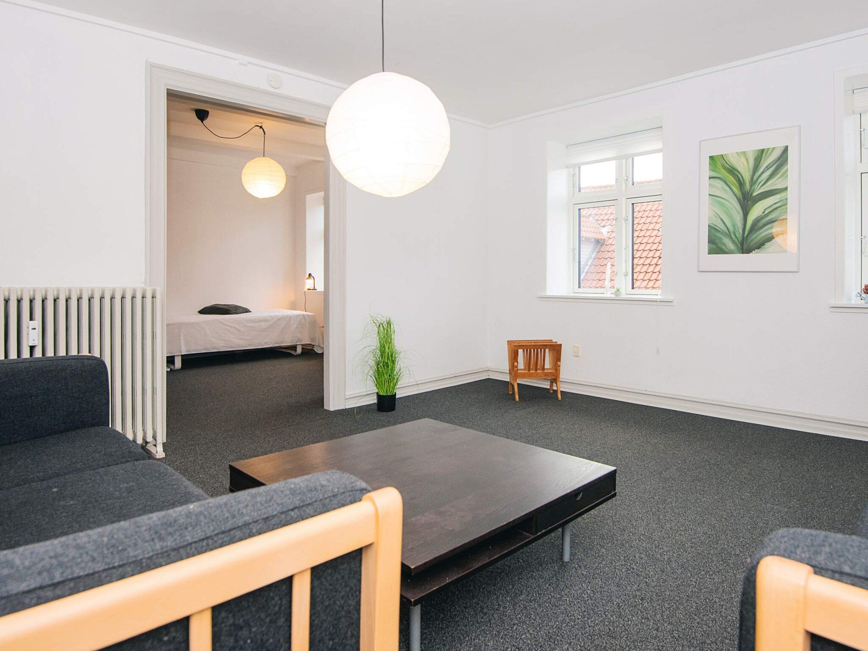A modern, well-lit room with a sofa, a coffee table, a small wooden chair, a potted plant, and a bed in the background.