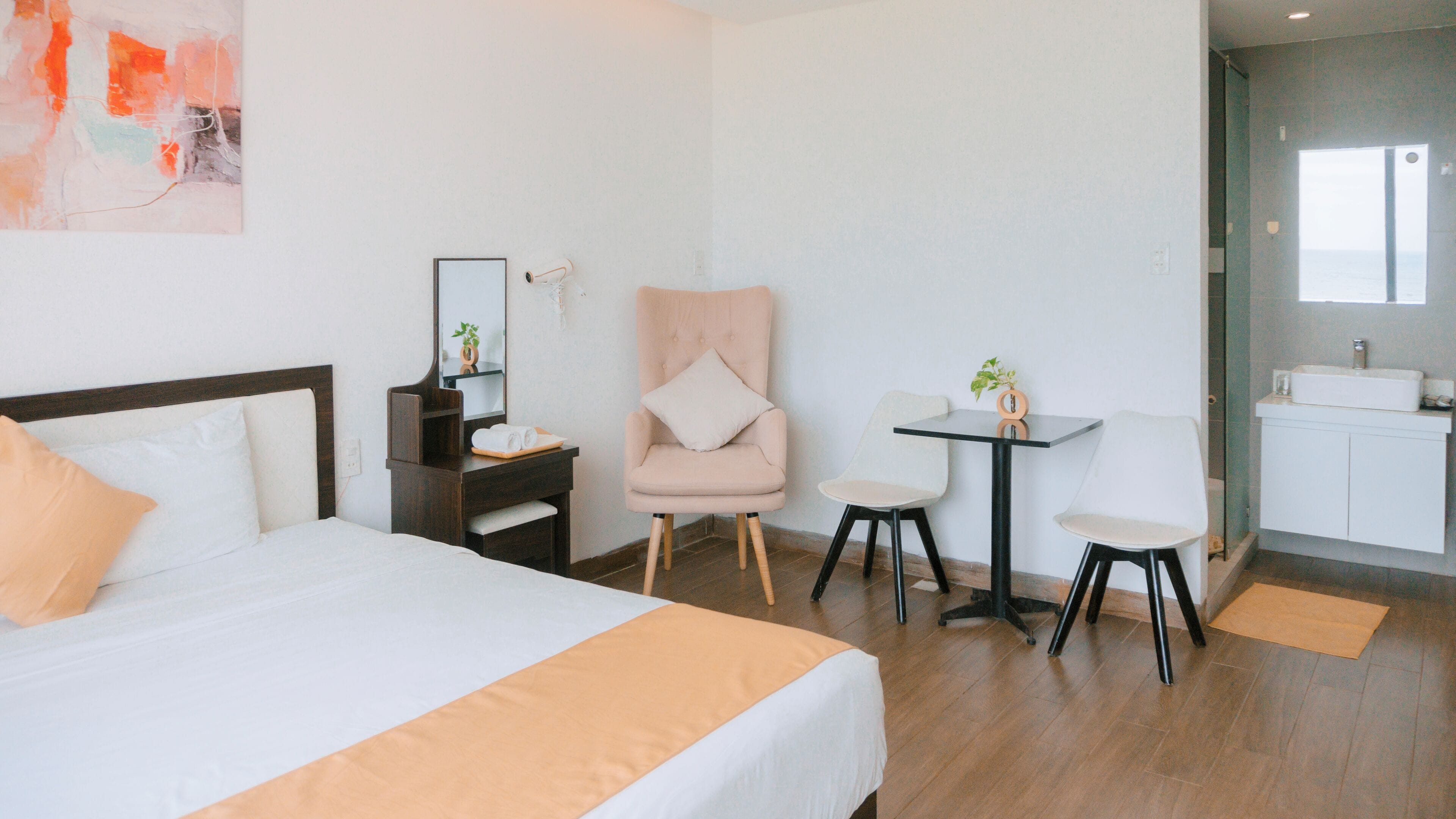 A hotel room with a bed, a desk with a chair, a small table with a chair, a mirror, and a bathroom with a sink.