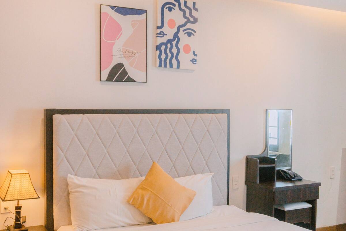 A neatly made bed with a quilted headboard, a bedside lamp, and two abstract wall art pieces.
