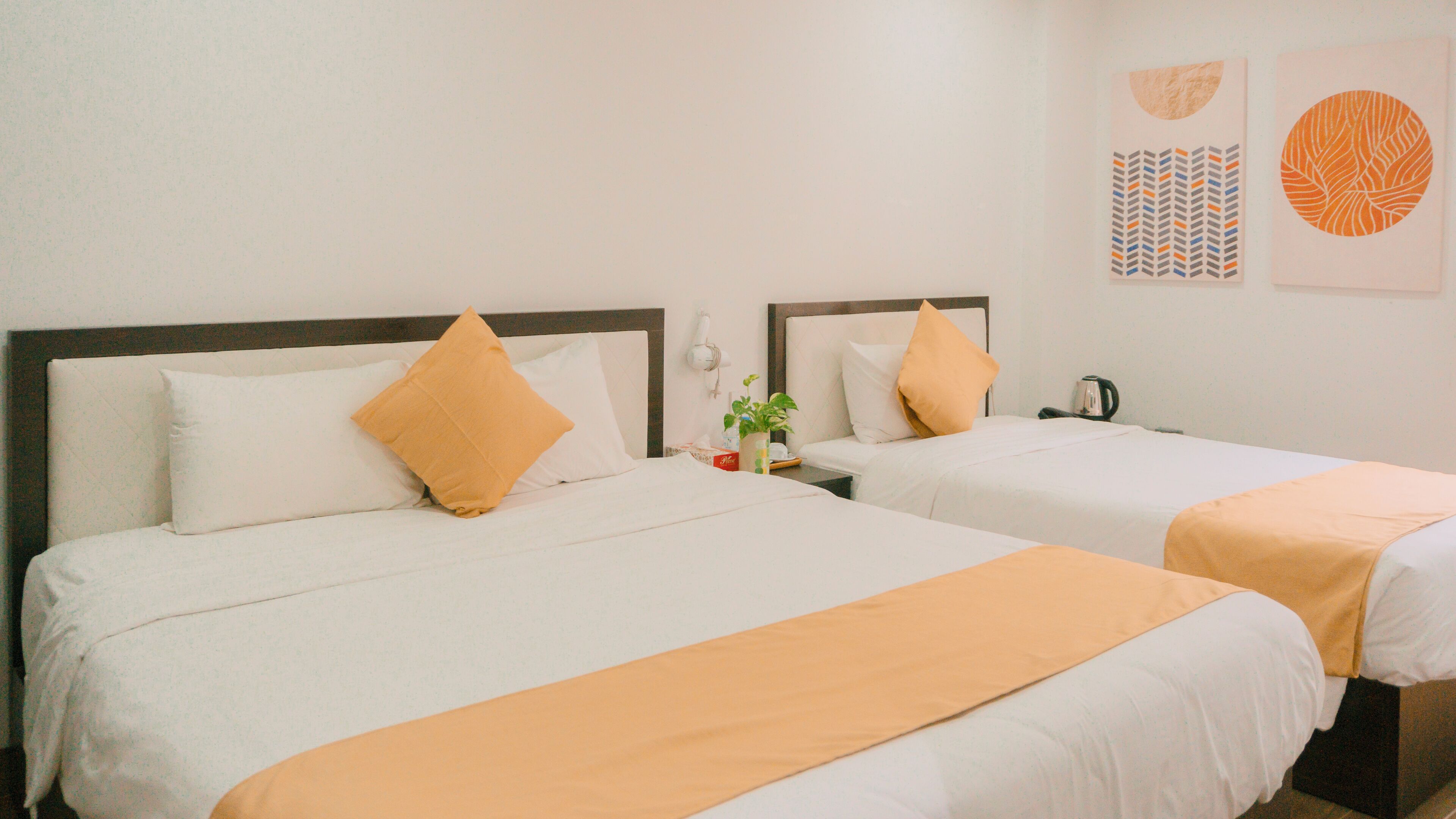 Two beds with white linens and orange accents, a bedside table with a plant, a kettle, and a wall-mounted phone.