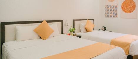 Two beds with white linens and orange accents, a bedside table with a plant, a kettle, and a wall-mounted phone.