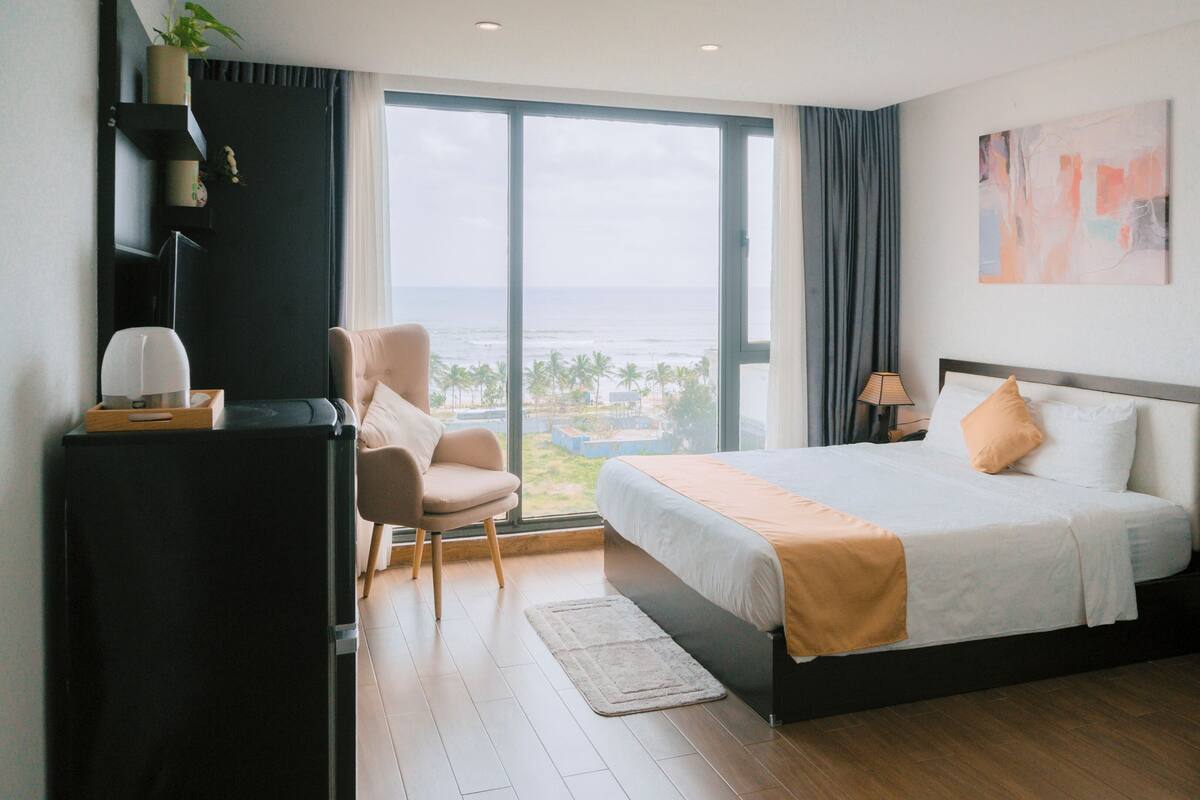 A hotel room with a large bed, a desk with a lamp, a chair, and a view of the ocean.