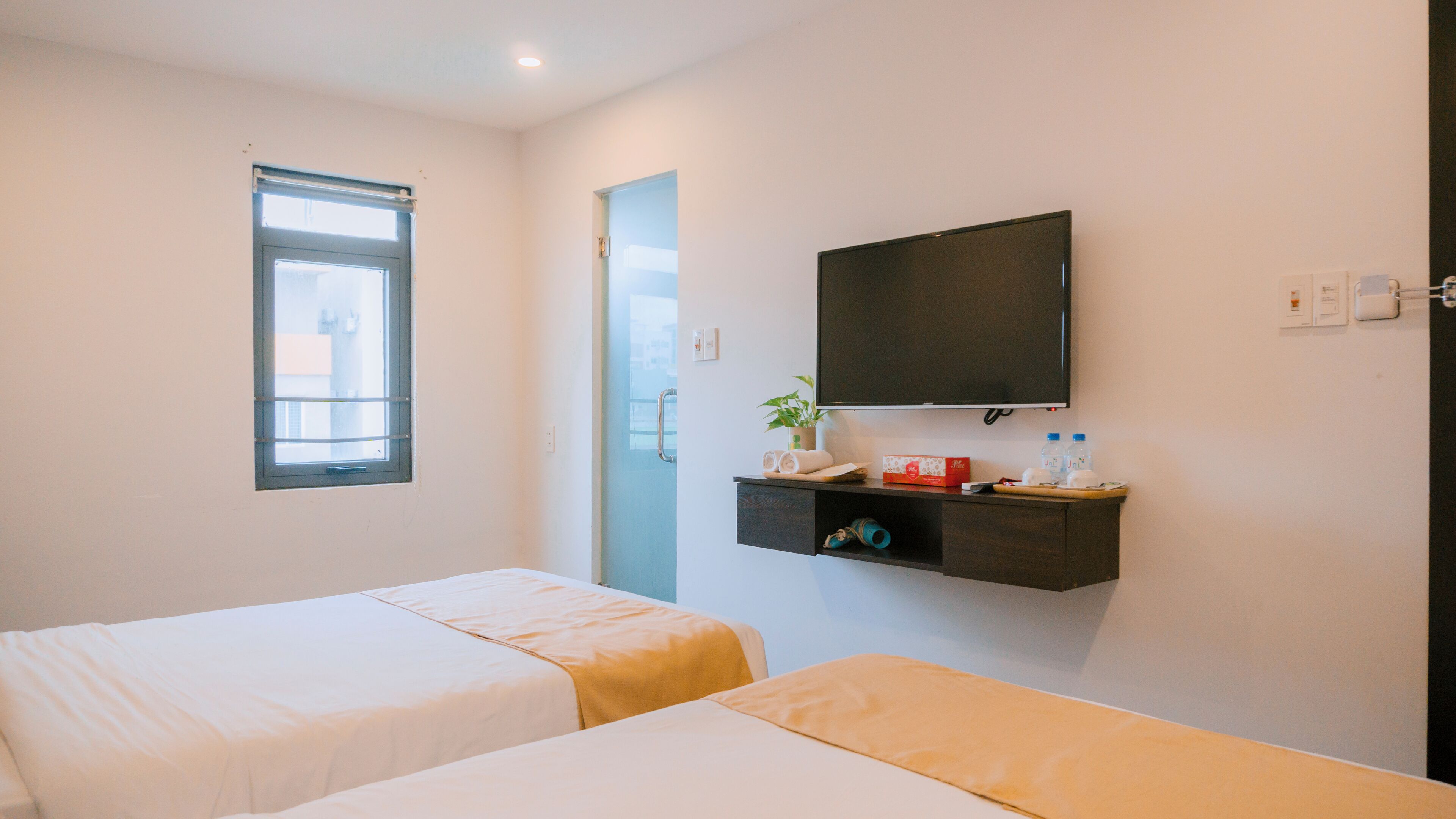 Habitación de hotel con dos camas, una ventana, un televisor de pantalla plana, un estante pequeño con artículos de aseo y una puerta con manija.