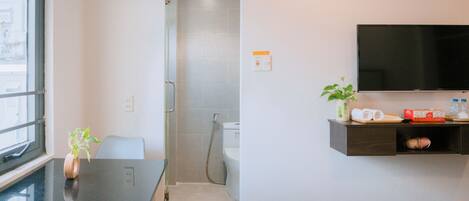 A hotel bathroom with a shower, toilet, and sink. There is a TV mounted on the wall, a small table with a plant, and a shelf with toiletries.