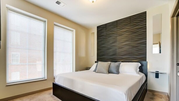A bedroom with a large bed, two windows, a wall-mounted headboard, and a door.