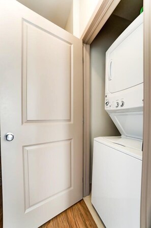 A laundry room with a door, a stacked washer and dryer, and a wooden floor.