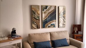 A living room with a beige sofa, two blue cushions, a coffee machine, and a white cabinet. There are three abstract paintings on the wall. - Ortigia Sweet Home in Syracuse (Syracuse)