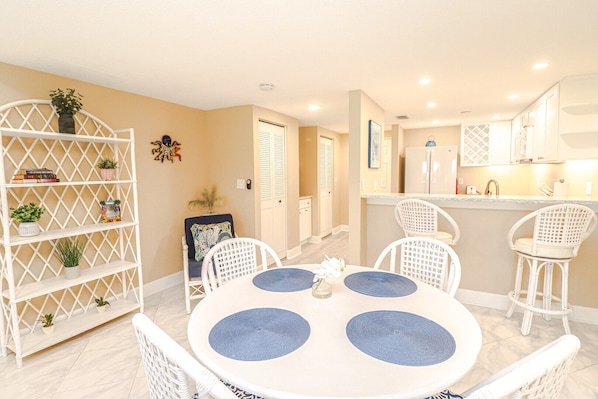A dining area with a round table, white chairs, and a white shelving unit with plants.