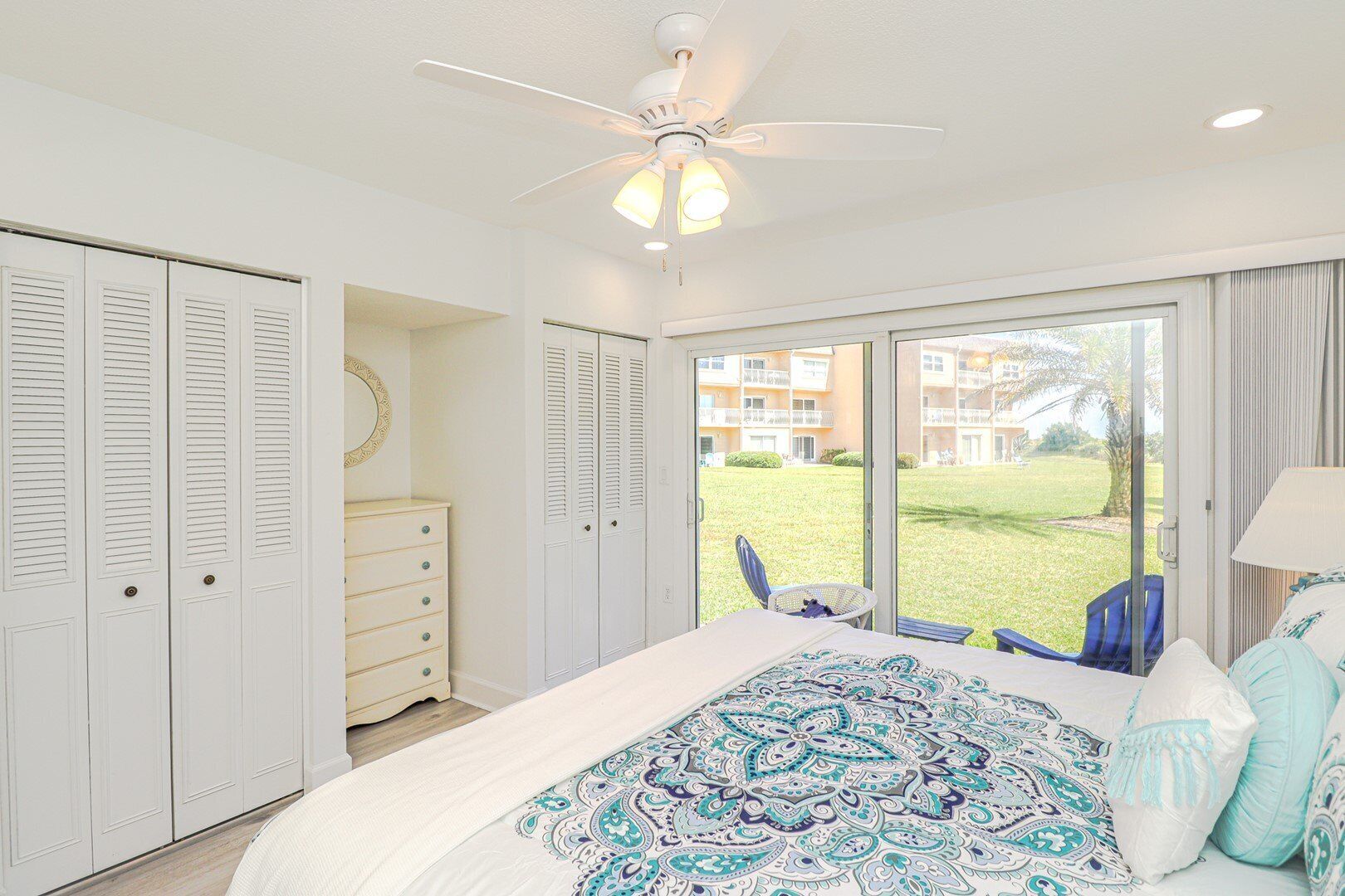 A bedroom with a bed, a ceiling fan, a dresser, and a view of a grassy area through a sliding glass door.