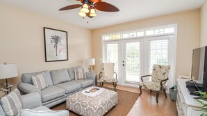 A living room with a grey sofa, patterned ottoman, and a ceiling fan.
