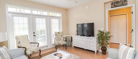 Un salon avec un ventilateur de plafond, un téléviseur posé sur un meuble blanc, deux fauteuils à motifs, un pouf à motifs et une plante en pot.