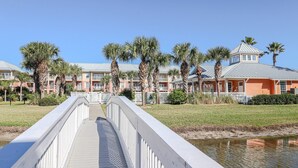 A wooden bridge leading to a hotel with multiple buildings, palm trees, and a body of water.