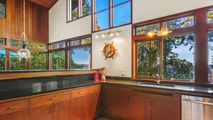 A modern kitchen with wooden cabinets, a black countertop, a stainless steel dishwasher, and large windows offering a view of trees. - Puzzle Point Lodge in Poulsbo (Poulsbo)