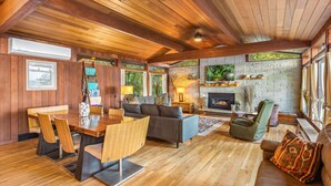 A spacious living area with a wooden ceiling, a fireplace, and a large window overlooking a garden. - Puzzle Point Lodge in Poulsbo (Poulsbo)