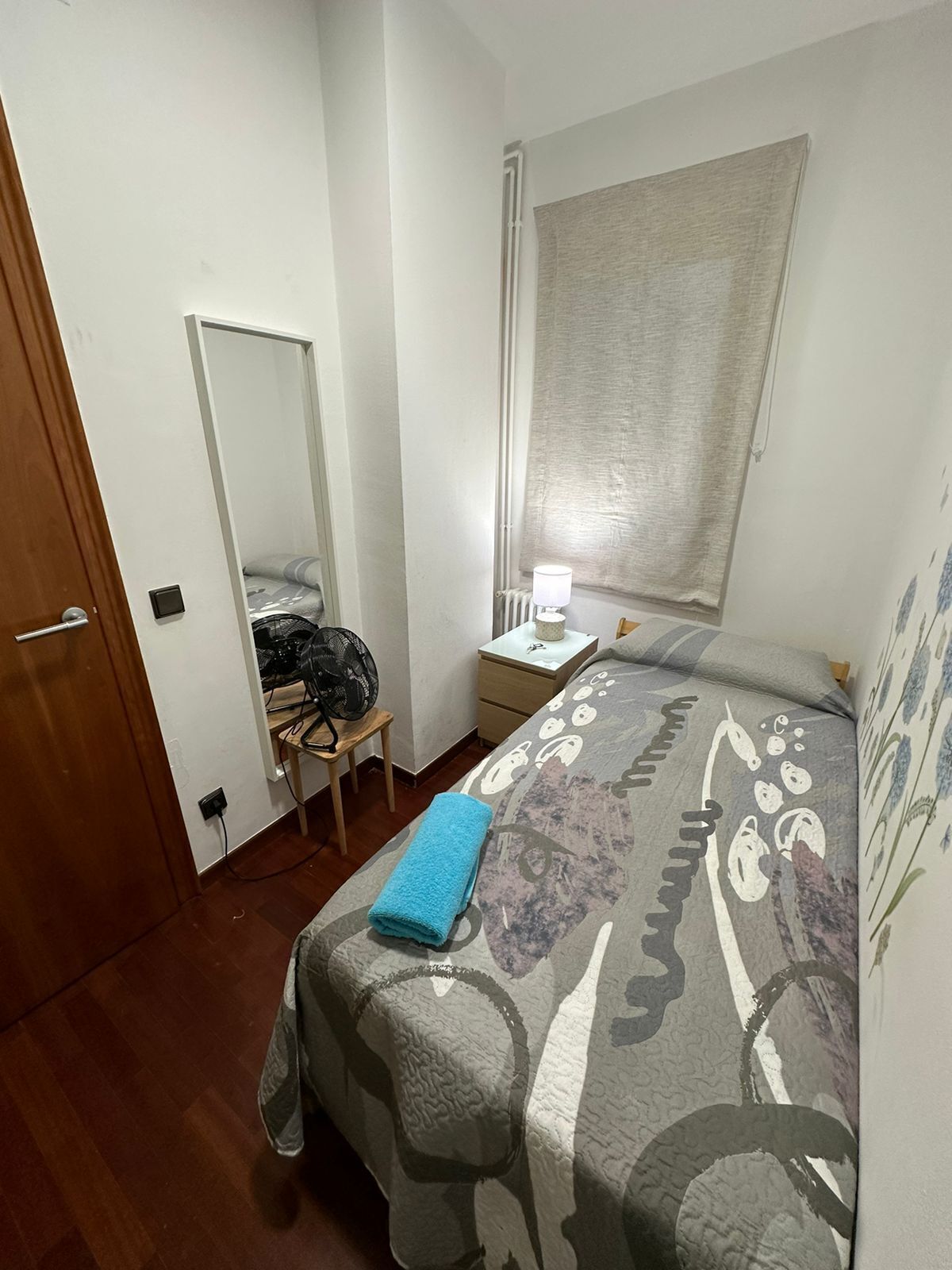 Een compact, netjes ingerichte slaapkamer met een bed, nachtkastje, spiegel en een raam met gordijn.