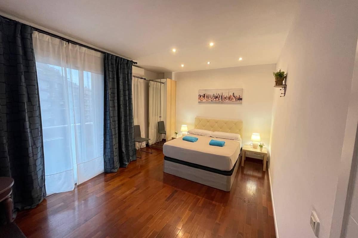 Habitación de hotel con una cama grande, vistas a la ciudad, una mesita de noche con lámpara y una silla.
