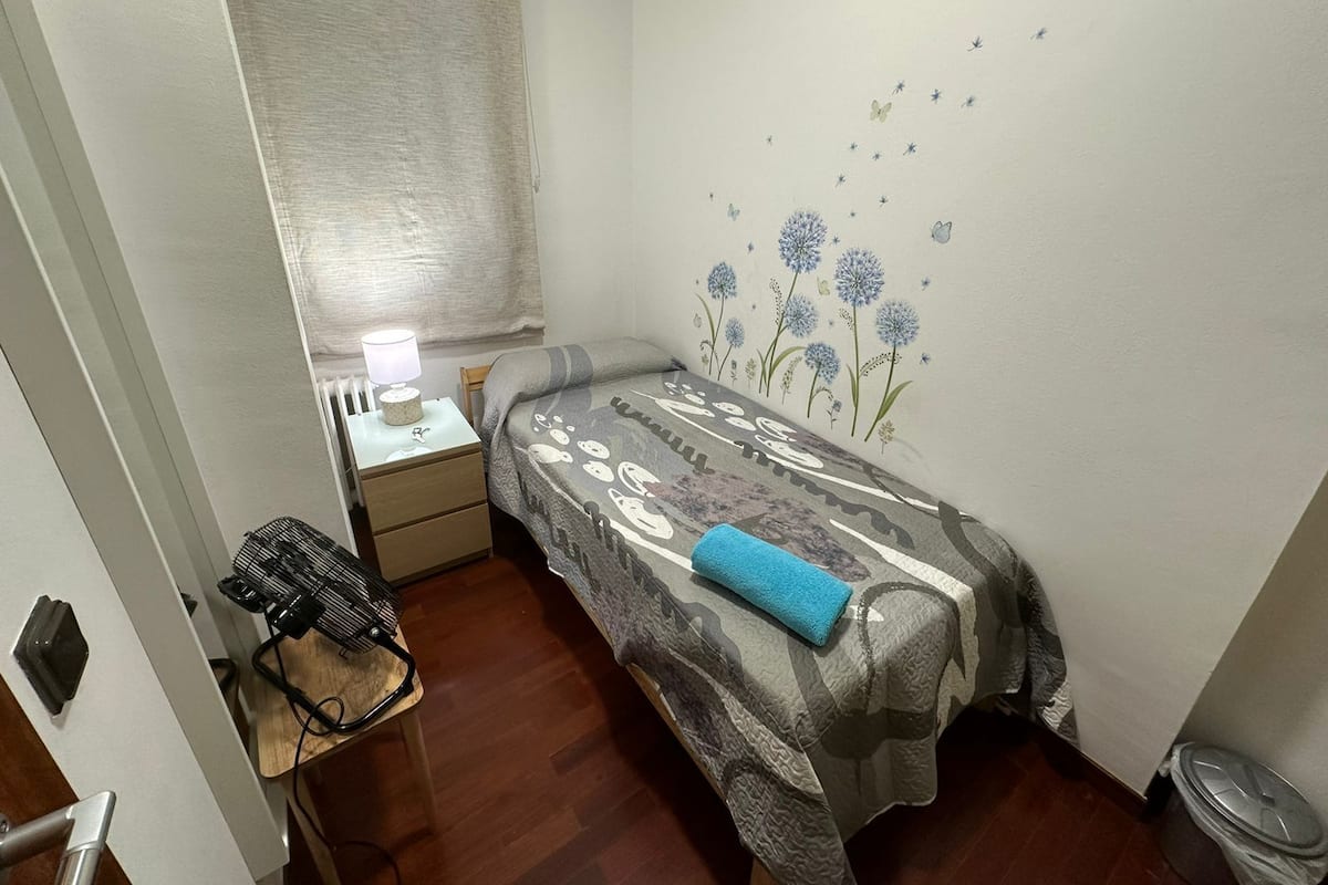 Un dormitorio pequeño y acogedor con una cama individual, una mesita de noche con lámpara, un radiador y una pared decorada con motivos florales.