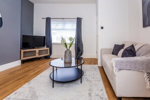 A modern living room with a flat-screen TV, a round glass coffee table, a beige sofa, and a decorative vase with flowers. - Toast Stays - The Garthhill (Ferryhill)