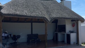 Un espacio al aire libre cubierto con techo de paja, zona de estar con sillas, una mesita y una parrilla.