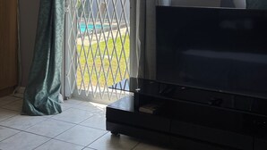 A living room with a flat-screen TV, a ceiling fan, and a view through a window with a metal gate. - Zoe & Saint (Cape Town)