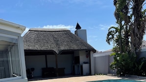 A backyard with a swimming pool, a thatched-roof structure, and a grassy area with scattered stones. - Zoe & Saint (Cape Town)