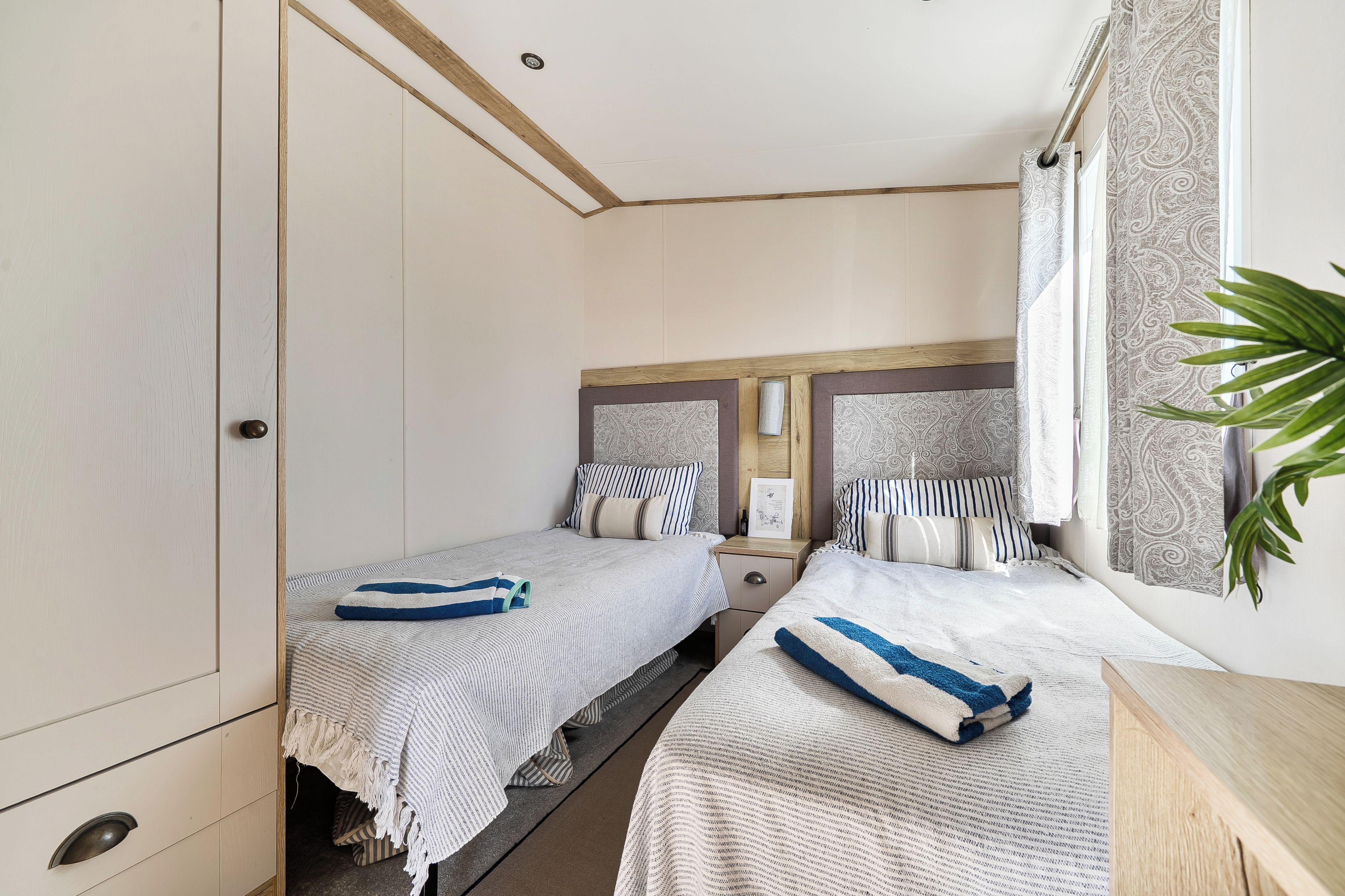 A bedroom with two beds, a wardrobe, a bedside table, and a window with curtains.