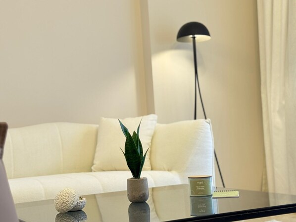 A modern living room with a white sofa, a small potted plant, a glass table with a candle, and a floor lamp. - Mynt Stay zed west Residence (Sheikh Zayed City)