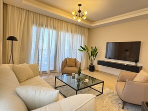 A modern living room with a sofa, armchair, coffee table, and a TV mounted on the wall. - Mynt Stay zed west Residence (Sheikh Zayed City)