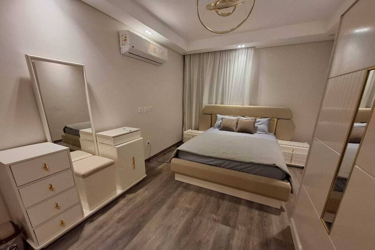 A bedroom with a bed, bedside tables, a dresser, a mirror, and an air conditioning unit.