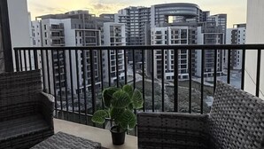 A balcony with wicker furniture and a potted plant, overlooking modern buildings. - Mynt Stay zed west Residence (Sheikh Zayed City)