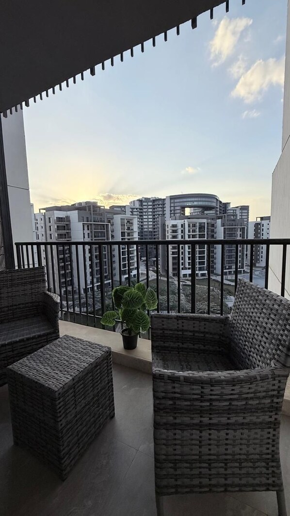 A balcony with wicker furniture and a potted plant, overlooking modern buildings. - Mynt Stay zed west Residence (Sheikh Zayed City)