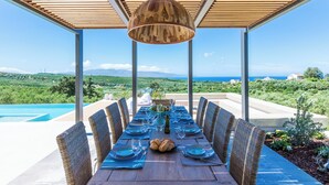 A dining table set for six with wicker chairs, a wooden pergola overhead, and a view of the sea and hills.