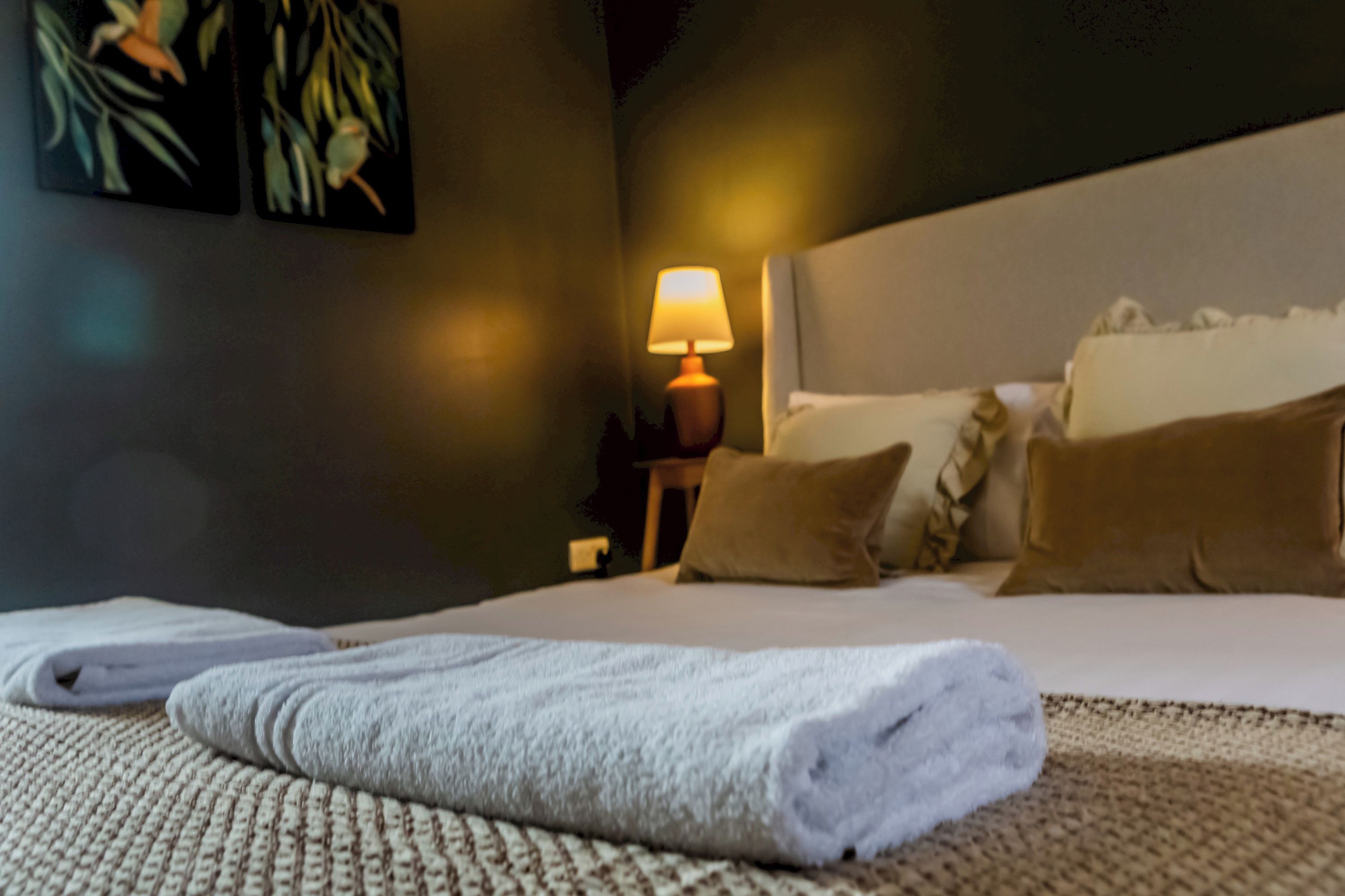 A neatly made bed with a folded towel, a bedside lamp, and a wall with a painting.