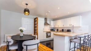 A modern kitchen with white cabinets, a central island with bar stools, a round dining table with chairs, and a hanging light fixture. - Hallgarth Street  (Sherburn Village)