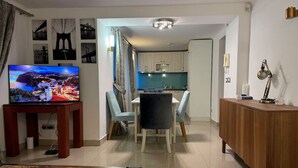 Un salon moderne comprenant un téléviseur, une table à manger et des chaises. Il y a également un coin cuisine avec des placards et un plan de travail.
