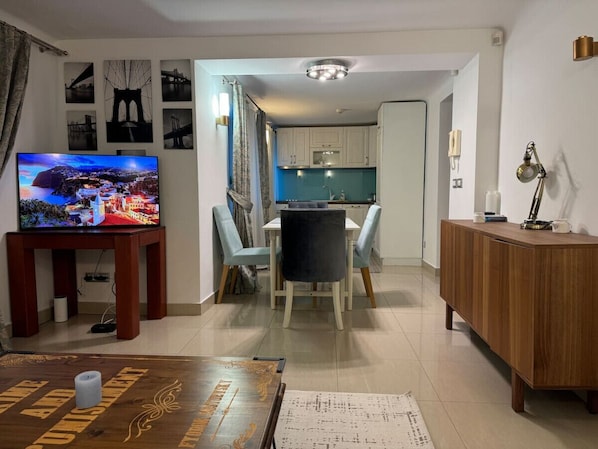 A modern living room with a TV, dining table, and chairs. There is a kitchen area with cabinets and a countertop.