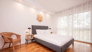 Une chambre avec un grand lit, un fauteuil en osier, une table de chevet avec des lampes, et une vue sur les arbres à travers des rideaux légers.