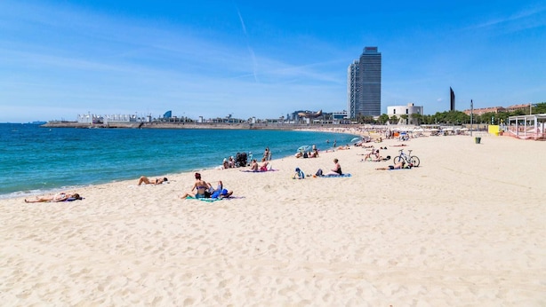 Une plage où des gens se prélassent au soleil et se baignent, un grand immeuble en arrière-plan et un ciel bleu dégagé.