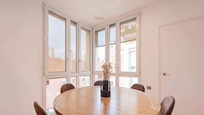 Une table ronde en bois, entourée de six chaises, un vase avec des fleurs séchées et de grandes fenêtres donnant sur la ville.