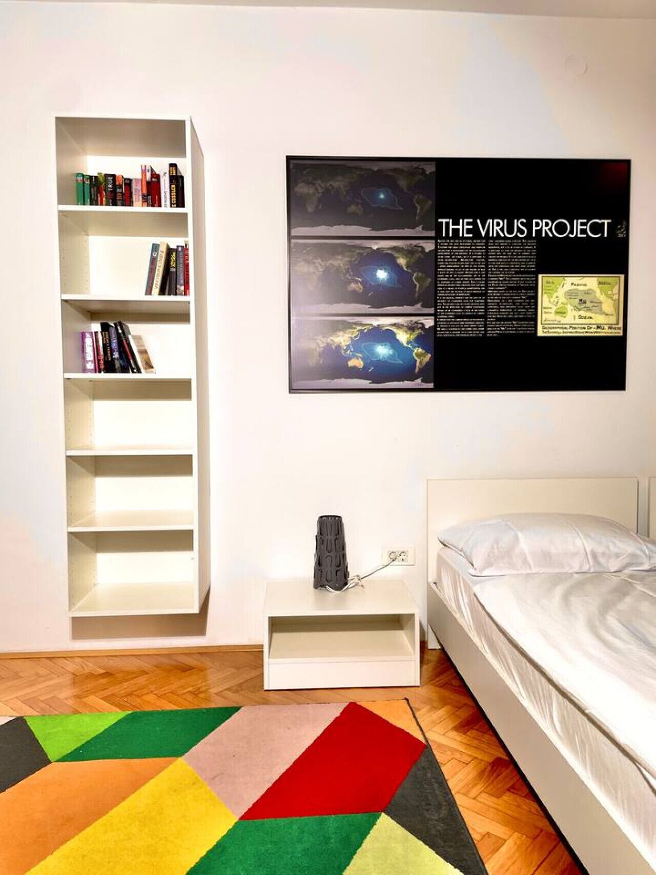 Een slaapkamer met een wit bed, een boekenkast, een ingelijste poster en een kleurrijk vloerkleed.