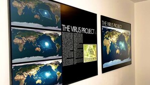 Ein Zimmer mit einem Bett, einem Nachttisch und einem an der Wand hängenden Poster mit dem Titel "THE VIRUS PROJECT", das Weltkarten zeigt.