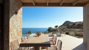 Eine Terrasse mit Holztisch und Stühlen, mit Blick auf einen Pool und das Meer.