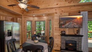 Ein gemütliches Blockhaus-Interieur mit Essbereich, Kamin und Fernseher, der eine Berglandschaft zeigt.