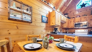A wooden cabin interior with a dining table set for four, wooden cabinets, a microwave, and a window with a star decoration. - Eagles Ridge Cabin · Hot Tub, Jacuzzi & Pool Table (Pigeon Forge)