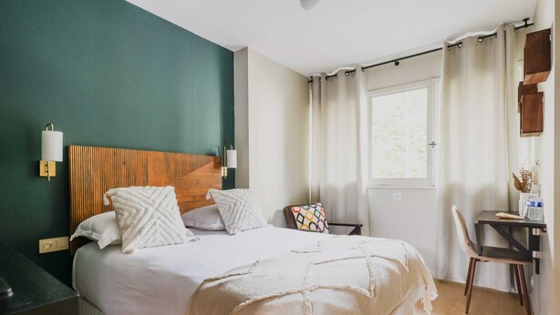 Quarto bem arrumado com cama, mesa de cabeceira, cadeira e janela com cortinas.