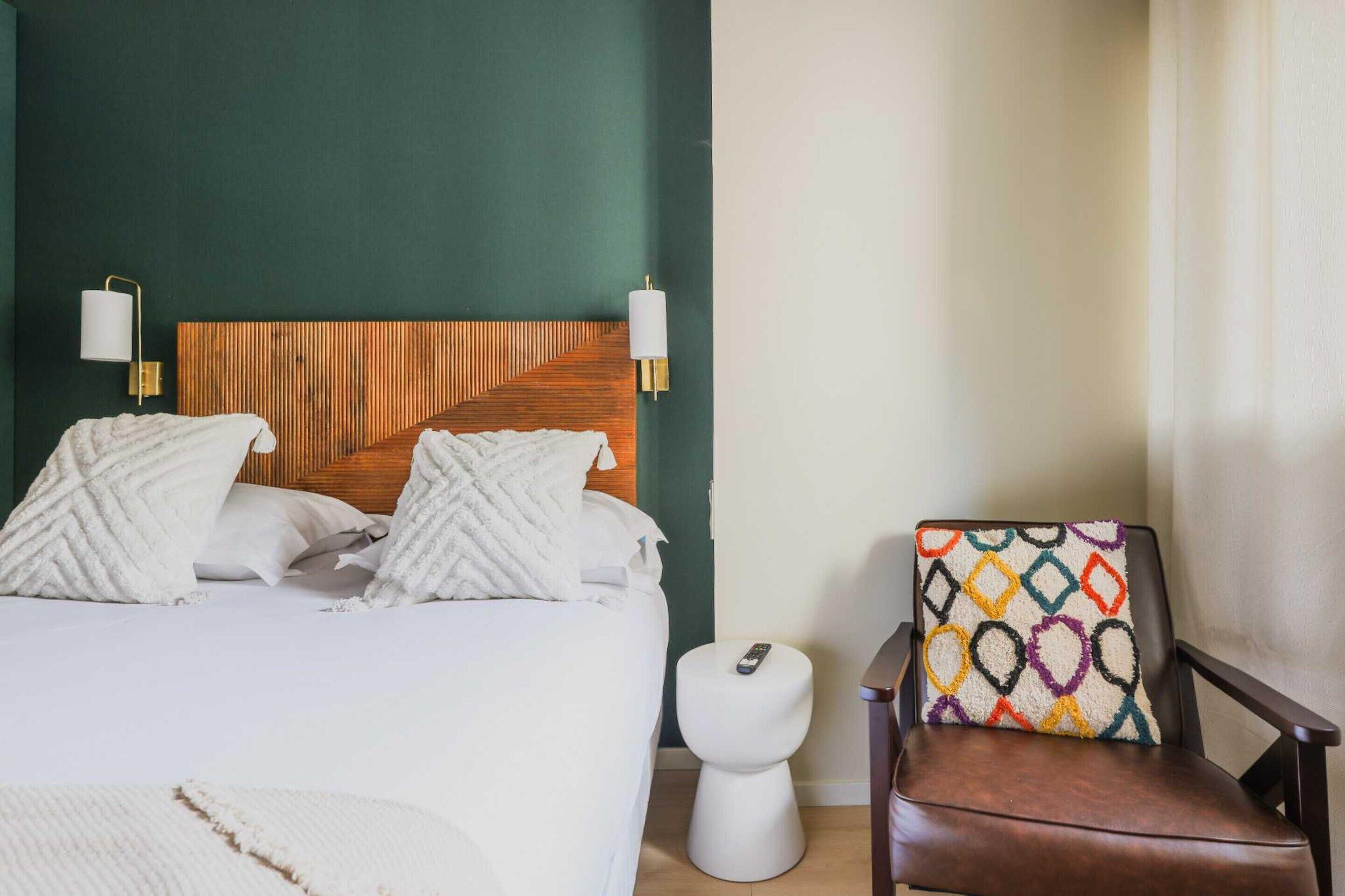 Habitación de hotel con cama, cabecera de madera, mesita de noche blanca, inodoro blanco y sillón de cuero marrón con cojín colorido.