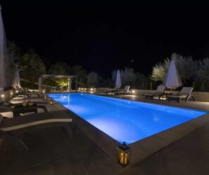 Una piscina con sillas de descanso y sombrillas, rodeada de árboles y una zona de estar techada.